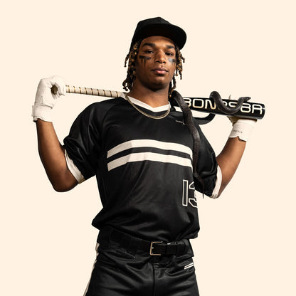 A baseball player in a black uniform and cap confidently holds the Warstic 2025 Bonesaber Hybrid Black Cobra (-3) BBCOR Bat (MBBSRHB25BK3) over his shoulders, with eye black, white gloves, and a plain light background.