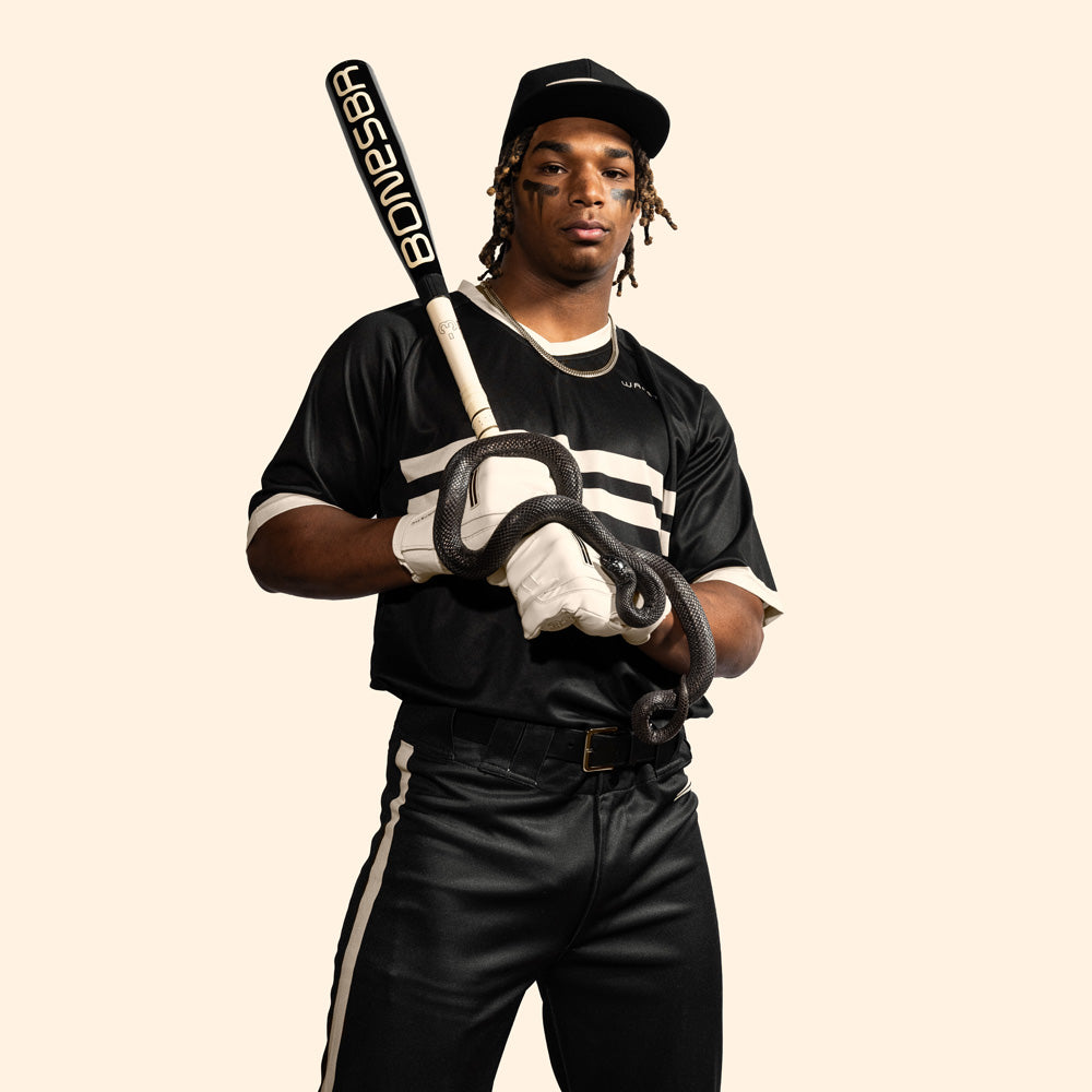 A baseball player in a black and white uniform grips the Warstic 2025 Bonesaber Hybrid Black Cobra (-3) BBCOR Bat (MBBSRHB25BK3), featuring a striking black snake design, standing confidently against a plain light background.