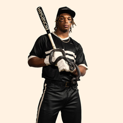 A baseball player in a black and white uniform grips the Warstic 2025 Bonesaber Hybrid Black Cobra (-3) BBCOR Bat (MBBSRHB25BK3), featuring a striking black snake design, standing confidently against a plain light background.