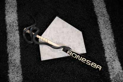 A black cobra coils around a Warstic 2025 Bonesaber Hybrid Black Cobra (-3) BBCOR Baseball Bat (MBBSRHB25BK3), which rests on home plate on a black turf field with white lines.