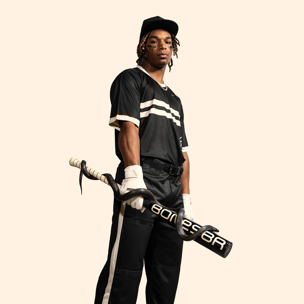 A baseball player in a black uniform and cap holds the Warstic 2025 Bonesaber Hybrid Black Cobra (-3) BBCOR Bat (MBBSRHB25BK3), wrapped with white tape, wearing white gloves and confidently facing the camera against a plain light background.