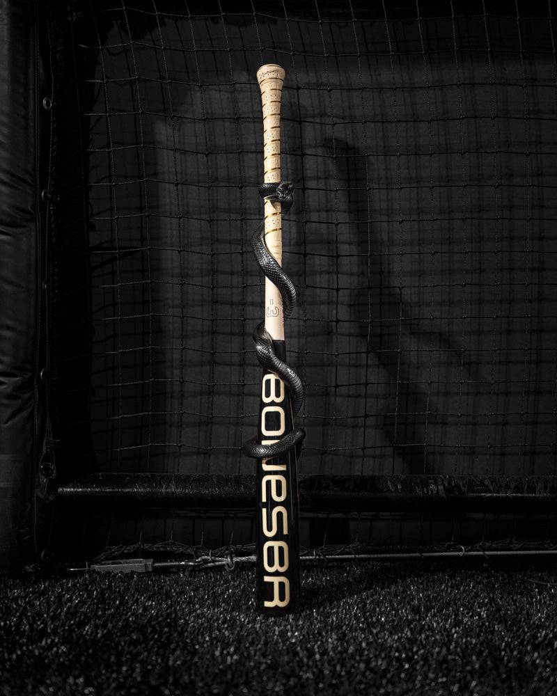 A Warstic 2025 Bonesaber Hybrid Black Cobra (-3) BBCOR Baseball Bat with a Katana2X Alloy Barrel is displayed upright, encircled by a black snake, set against a netted indoor backdrop.