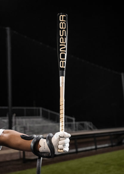 A person in a white glove holds the 2025 Warstic Bonesaber Hybrid Black Cobra (-3) BBCOR Baseball Bat (MBBSRHB25BK3) upright on a nighttime field, with empty bleachers and a black snake coiled around their arm.