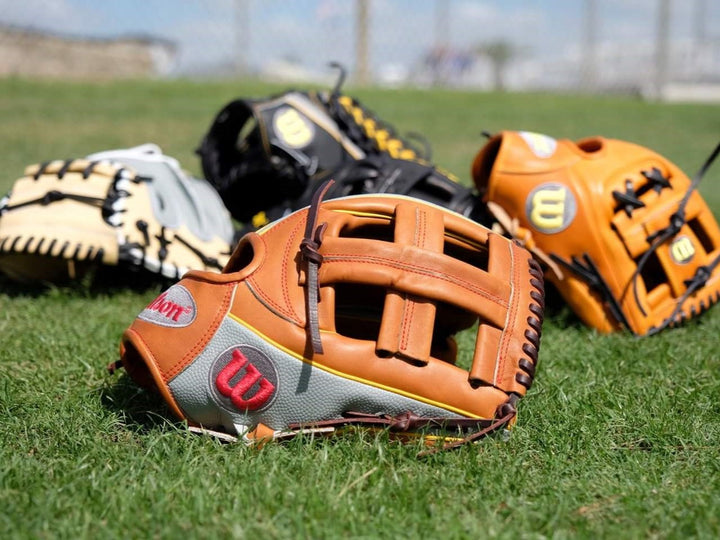 A close-up of four baseball gloves lying on green grass, with a metal fence and blurry background outdoors on a sunny day.