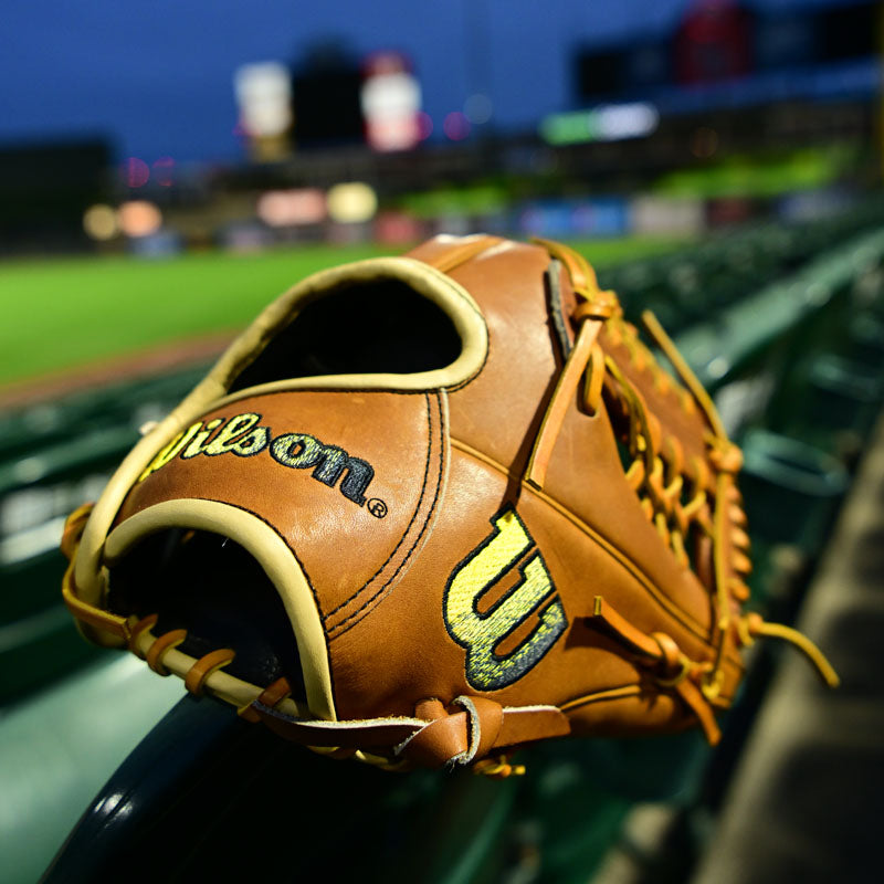 A brown Wilson baseball glove with yellow accents rests on a green stadium seat, with a baseball field and blurred scoreboard visible in the background at dusk.