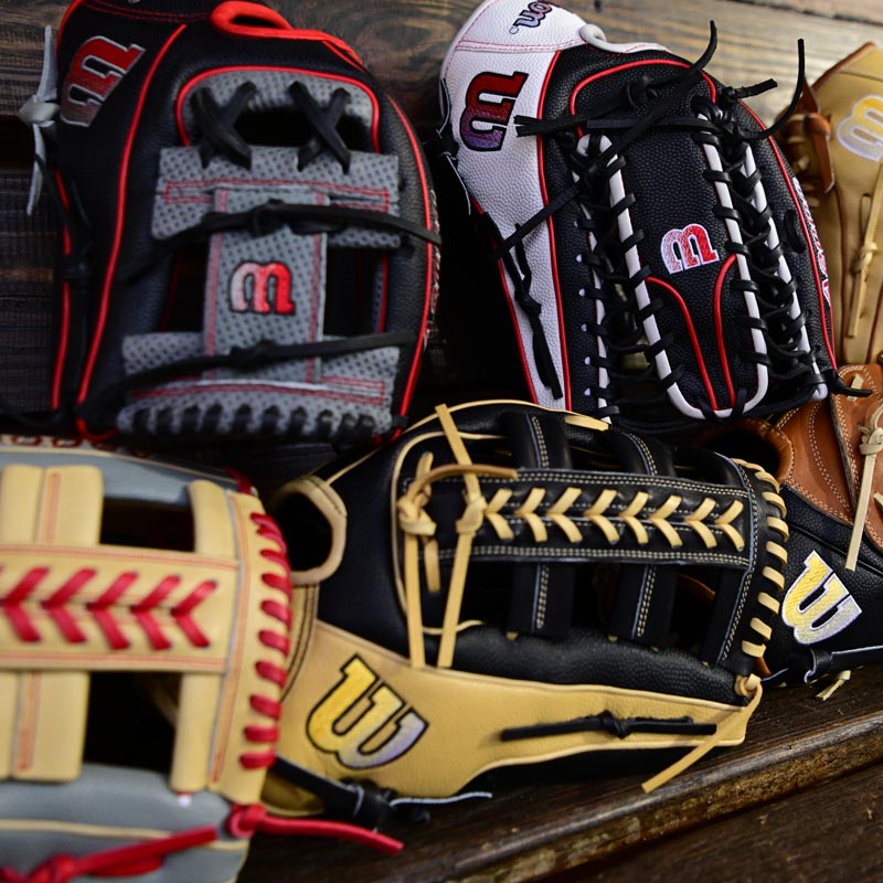 A close-up of several colorful baseball gloves with distinctive stitching and “W” logos, arranged overlapping each other on a wooden surface.