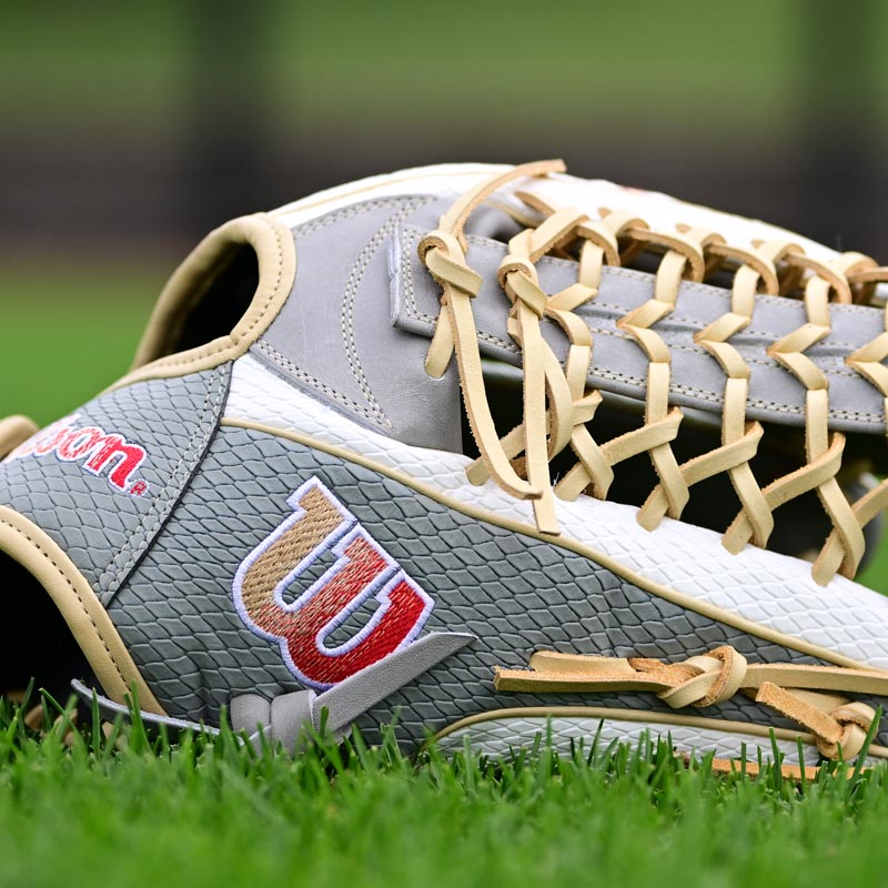 A close-up of a gray and white Wilson baseball glove with tan laces laying on green grass, showing the red and white Wilson logo prominently.