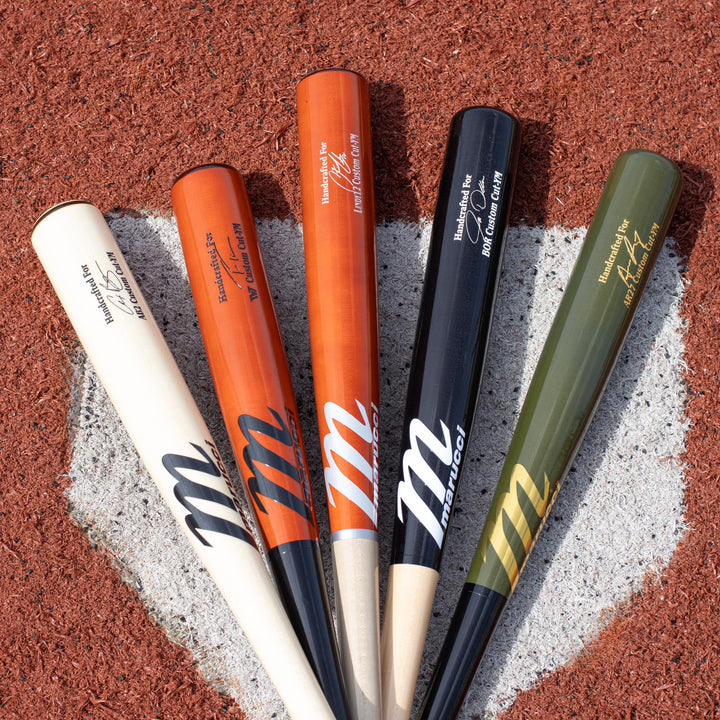 Five wooden baseball bats of different colors (white, orange, black, and green) are arranged fanned out on a brown dirt field, positioned over home plate. Each bat displays branding and model text.