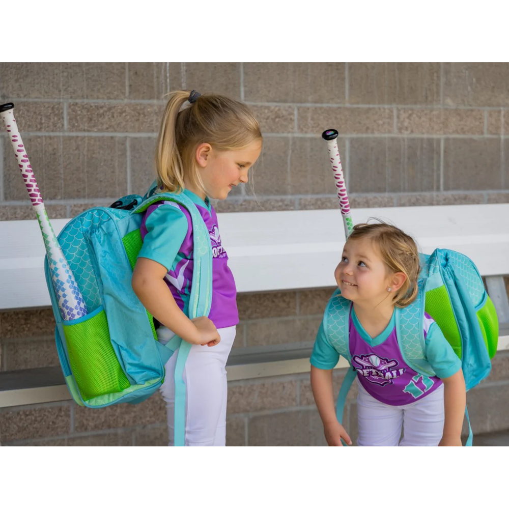 Two young girls in matching softball uniforms and backpacks, each holding a lightweight Rip It 2022 Spark -12 Fastpitch Softball Bat (SRPK), smile at each other while standing by a bench against a brick wall.