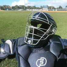 A Force3 Hockey Style Defender Catcher's Helmet (BD22) rests on the grass of a sunny baseball field, with palm trees and the Palm Beach Atlantic scoreboard in the background.