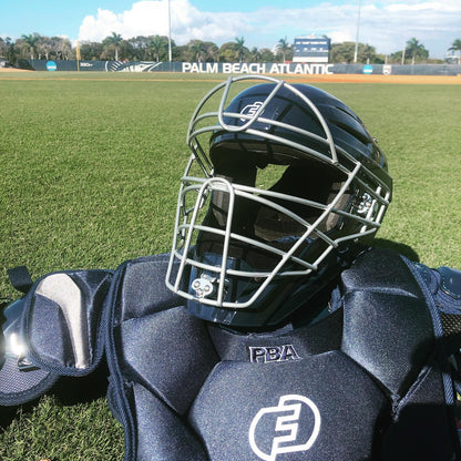 A Force3 Hockey Style Defender Catcher's Helmet (BD22) rests on the grass of a sunny baseball field, with palm trees and the Palm Beach Atlantic scoreboard in the background.