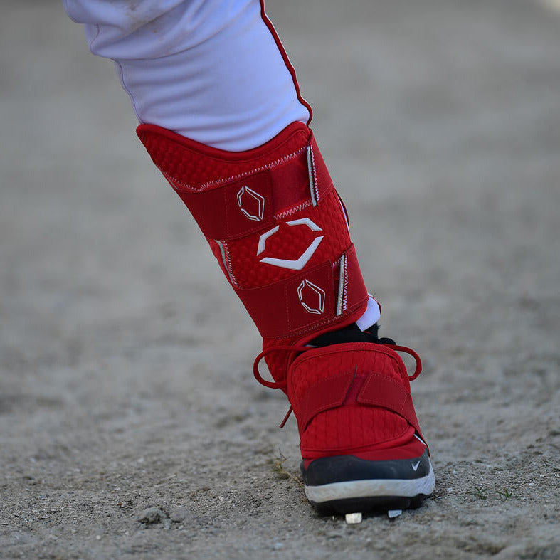 A baseball player's lower leg, dressed in white pants and a red cleat, is shown wearing the EvoShield PRO-SRZ 2.0 Batter's Leg Guard (WB5726901), standing on sandy ground.