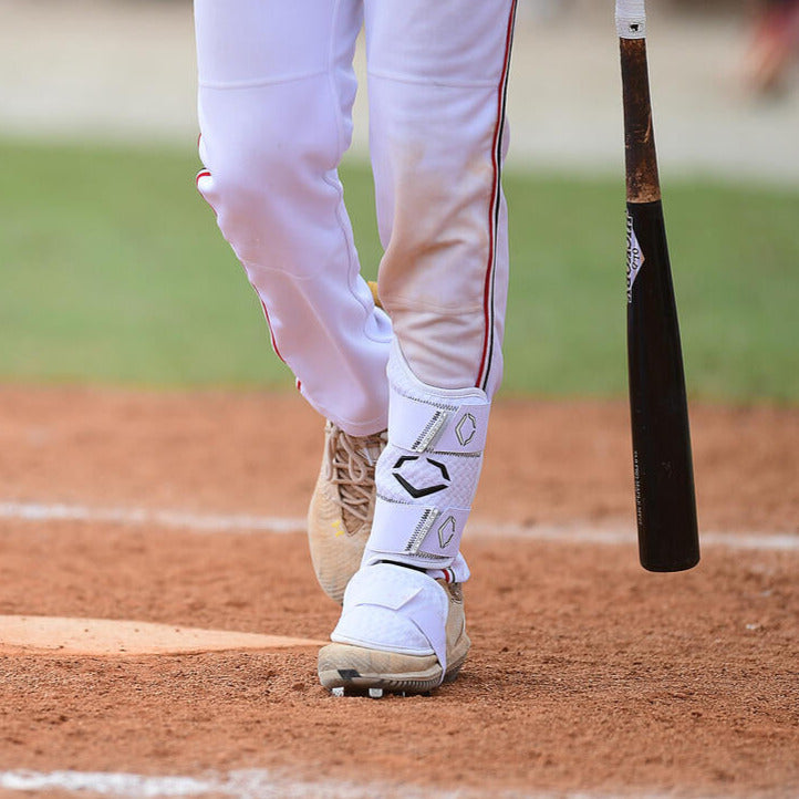 A baseball player, visible from the lower legs down, stands at home plate in white pants and an EvoShield PRO-SRZ 2.0 Batter’s Leg Guard (WB5726901), holding a black bat. The EvoShield brand is highlighted on the leg guard.