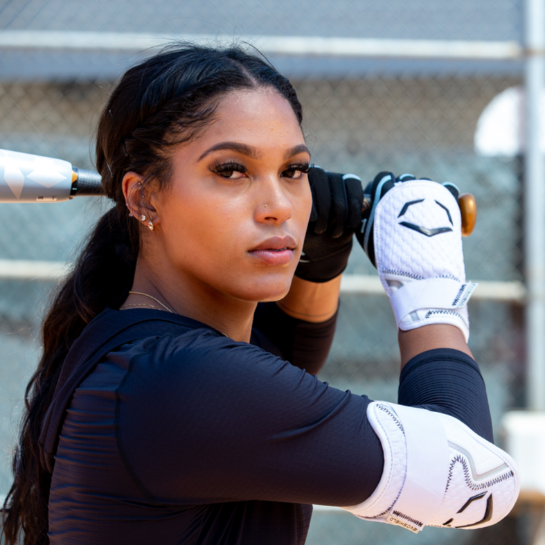 A woman in a black athletic outfit and white batting gloves wears the EvoShield PRO-SRZ 2.0 Batter's Hand Guard (WB5726801) for added protection as she holds a bat over her shoulder by a chain-link fence on a sunny day.