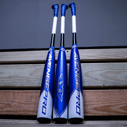 Three 2023 AXE Avenge Pro (-5) USSSA Baseball Bats with white grips stand upright side by side against wooden bleachers and a gray wall, highlighting their 3-piece composite design.