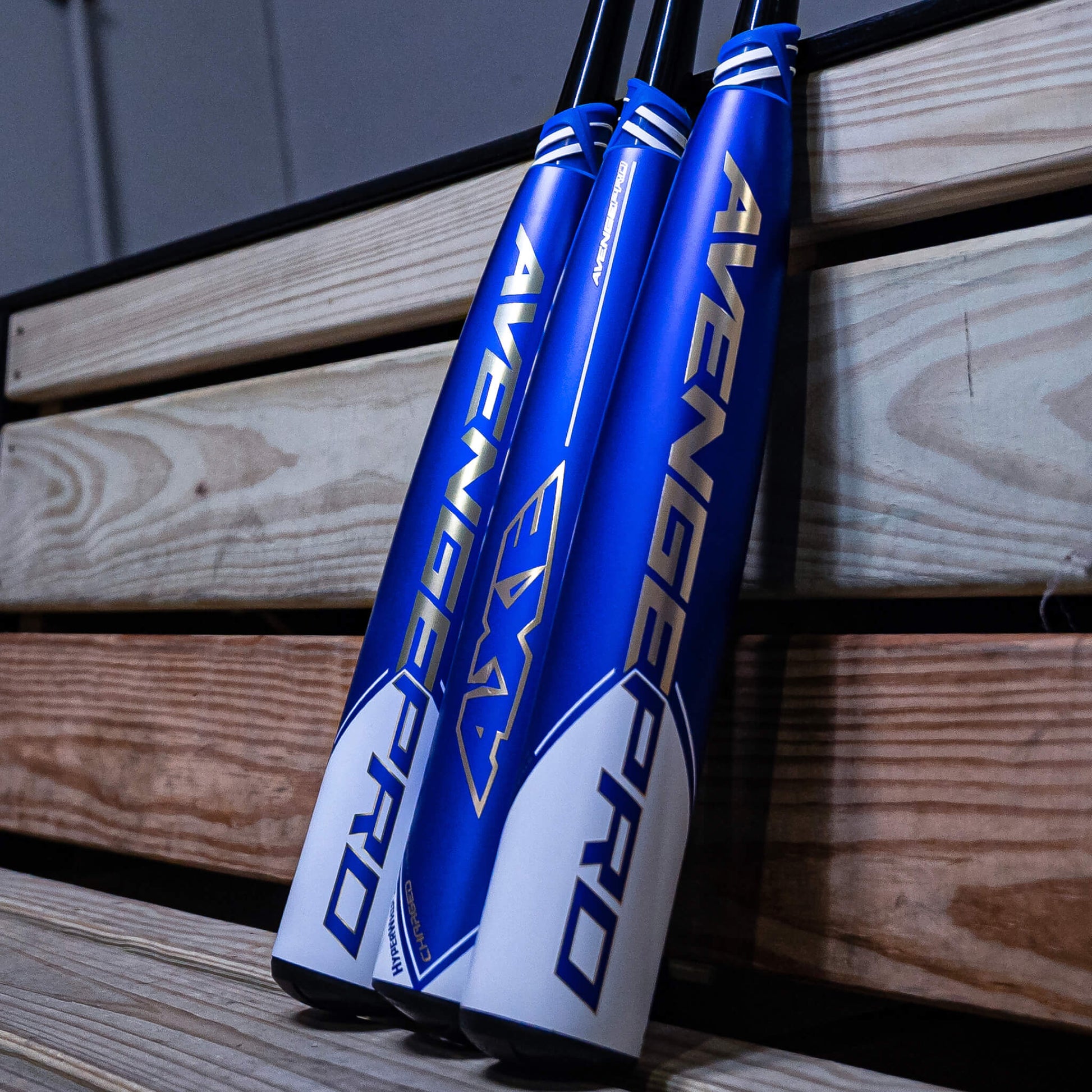 Three blue and white 2023 AXE Avenge Pro (-5) 2 5/8" USSSA Baseball Bats (USED) with gold lettering are leaning against wooden bleachers indoors.