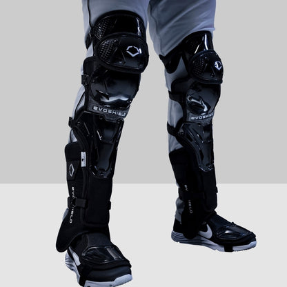 A person in gray pants and black Nike shoes stands on a light floor with a gray background, showcasing EvoShield Pro-SRZ Gel-to-Shell™ Catcher's Lower Leg Guards (WB570800/WB571030) for elite catcher’s protection.