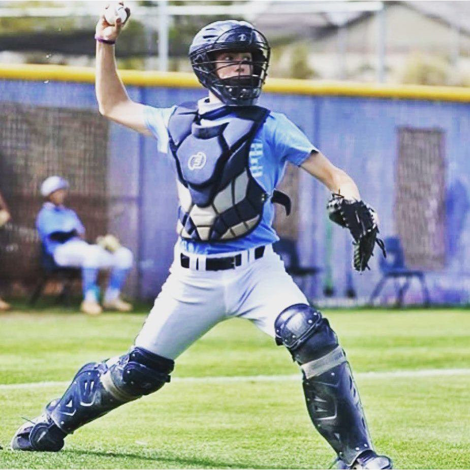 A young baseball catcher wearing Force3 Catcher's Leg Guards with DuPont Kevlar (BC8) is mid-throw on a grassy field, while another player sits in the background near a blue fence.