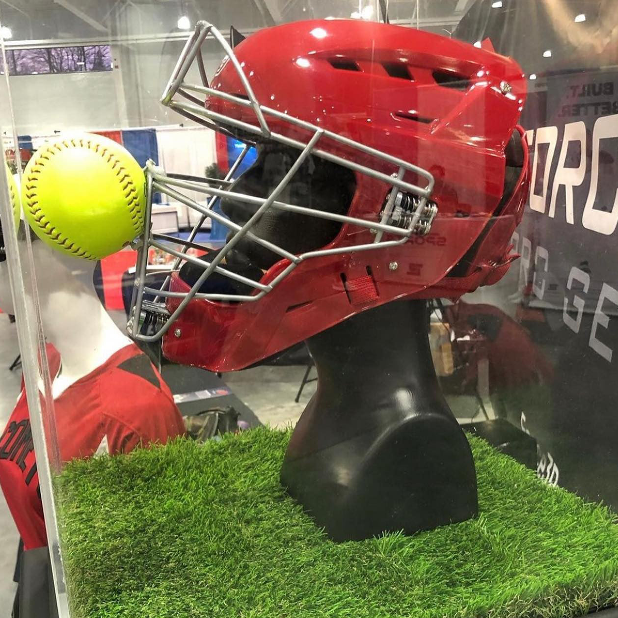 The Force3 Hockey Style Defender Catcher's Helmet: BD22, a red softball helmet with a face guard, is showcased on a mannequin head behind glass, with a yellow softball pressed to the guard atop artificial grass.