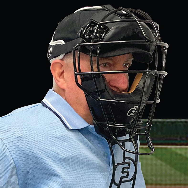 A person wearing a Force3 Pro Gear Traditional Defender Mask: BD31 by Force3 and a light blue collared shirt stands against a black background, with part of a sports field visible, emphasizing baseball safety.