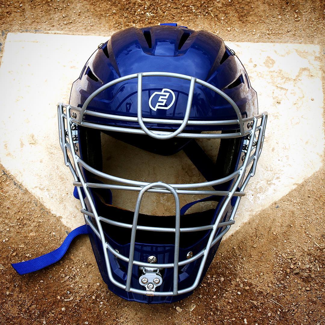 The Force3 Hockey Style Defender Catcher's Helmet: BD22, featuring a silver metal cage, rests on home plate, emphasizing umpire safety against the dirt of the baseball field.