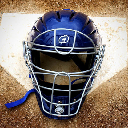The Force3 Hockey Style Defender Catcher's Helmet: BD22, featuring a silver metal cage, rests on home plate, emphasizing umpire safety against the dirt of the baseball field.