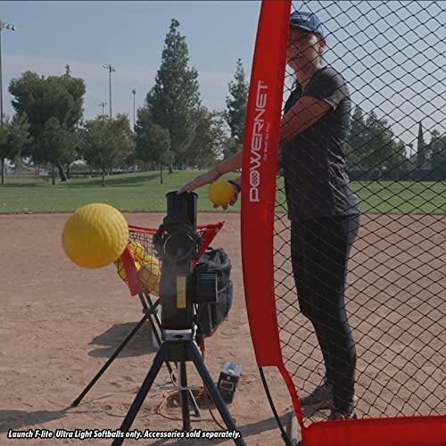 A person uses the PowerNet Launch F-Lite Pitching Machine with PowerNet Fastpitch Softballs (1194-2) on a baseball field, sending yellow softballs toward a net. Trees and light poles can be seen in the background.