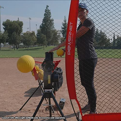 A person uses the PowerNet Launch F-Lite Pitching Machine with PowerNet Fastpitch Softballs (1194-2) on a baseball field, sending yellow softballs toward a net. Trees and light poles can be seen in the background.