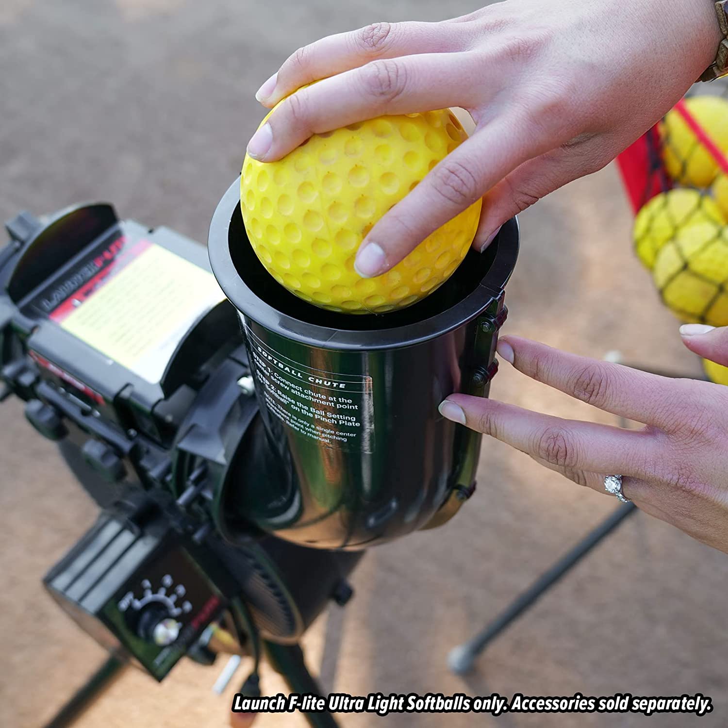 Outdoors, someone loads a yellow PowerNet Launch F-Lite Ultra Light Softball (1194-2) into a black PowerNet pitching machine. A bucket of softballs sits nearby. Only use Launch F-Lite Ultra Light Softballs. Accessories sold separately.