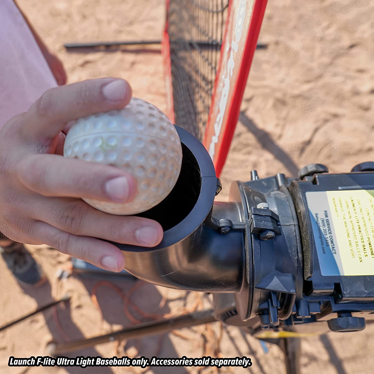 A person loads a white PowerNet Launch F-Lite Pitching Machine Baseball (1194-1) into a black machine on a sandy field. Text notes that PowerNet balls and other accessories are sold separately.