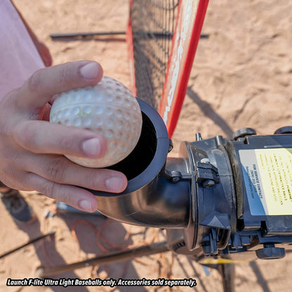 A person loads a white PowerNet Launch F-Lite Pitching Machine Baseball (1194-1) into a black machine on a sandy field. Text notes that PowerNet balls and other accessories are sold separately.