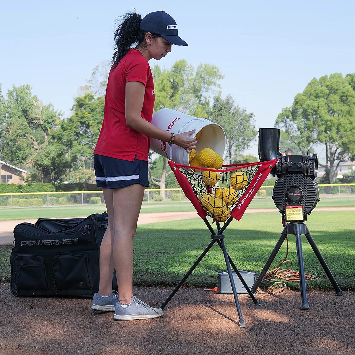 A woman in athletic wear pours PowerNet Launch F-Lite Pitching Machine Fastpitch Softballs (1194-2) into a red netted feeder by a PowerNet pitching machine on a baseball field, with her gear bag placed nearby.