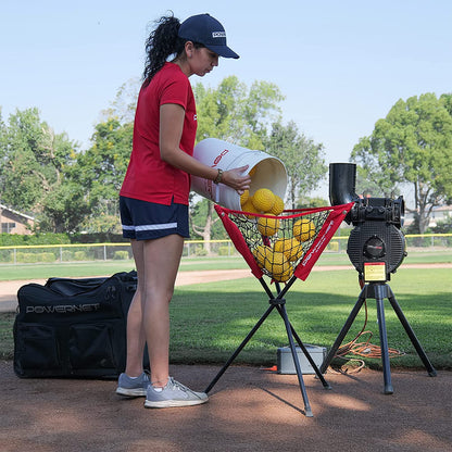 A woman in athletic wear pours PowerNet Launch F-Lite Pitching Machine Fastpitch Softballs (1194-2) into a red netted feeder by a PowerNet pitching machine on a baseball field, with her gear bag placed nearby.