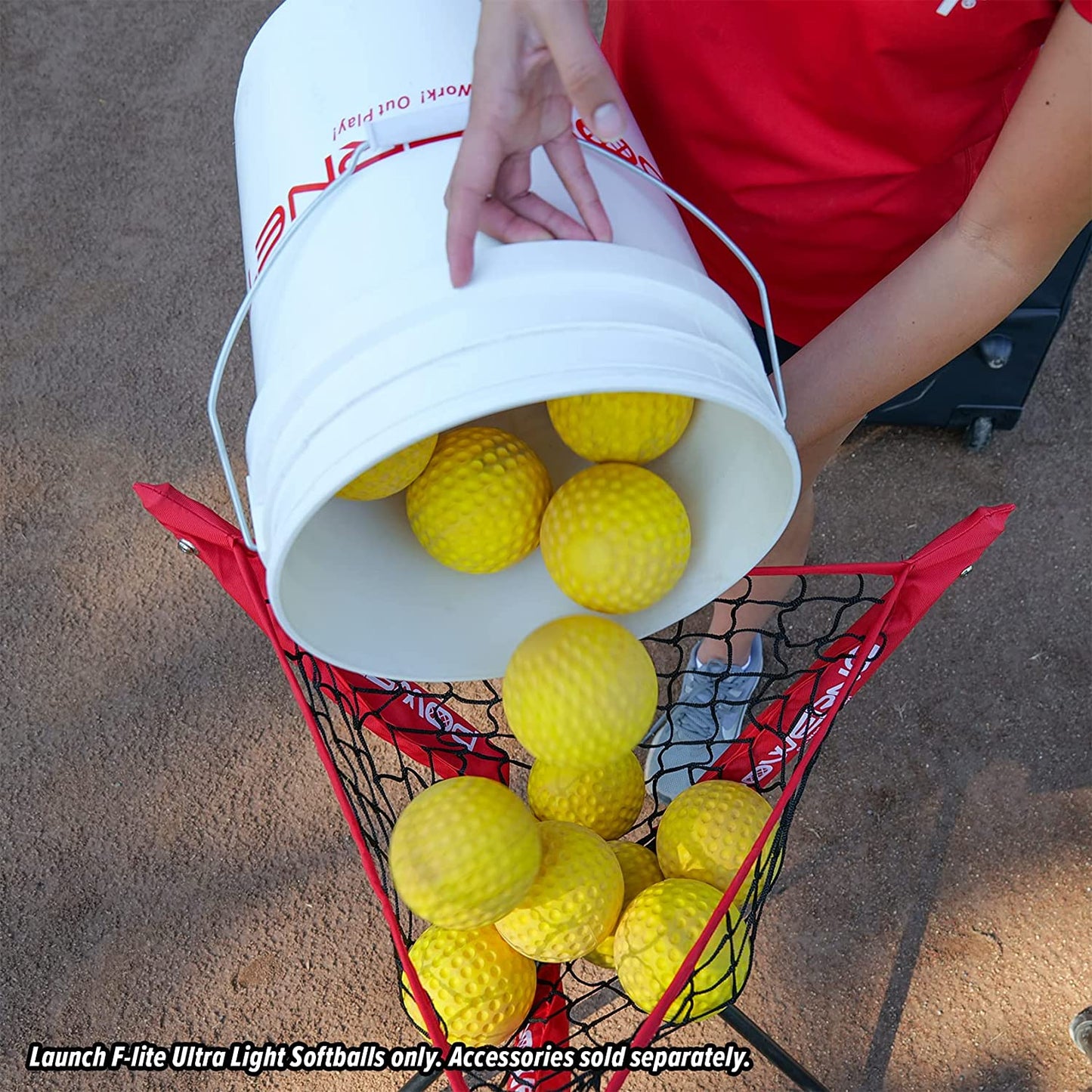 A person pours bright yellow PowerNet Launch F-Lite Pitching Machine Fastpitch Softballs: 1194-2 from a large white bucket into a black and red netted basket. Text reads: Use Launch F-Lite Ultra Light Softballs only. Accessories sold separately.