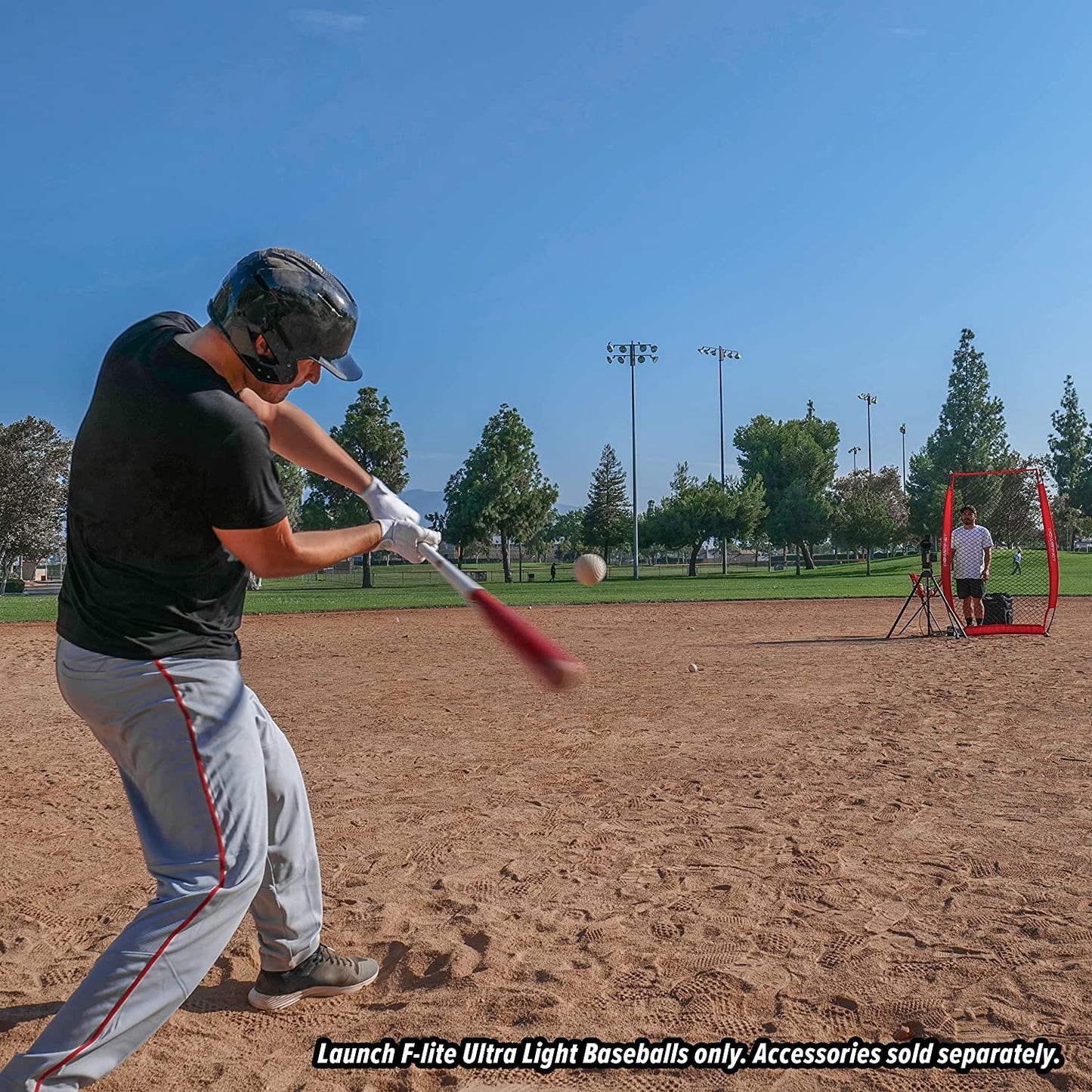 A player in a helmet hits PowerNet Launch F-Lite Pitching Machine Baseballs (1194-1) on a dirt field, while another person sits near a red PowerNet practice net. Trees border the field beneath a clear sky.