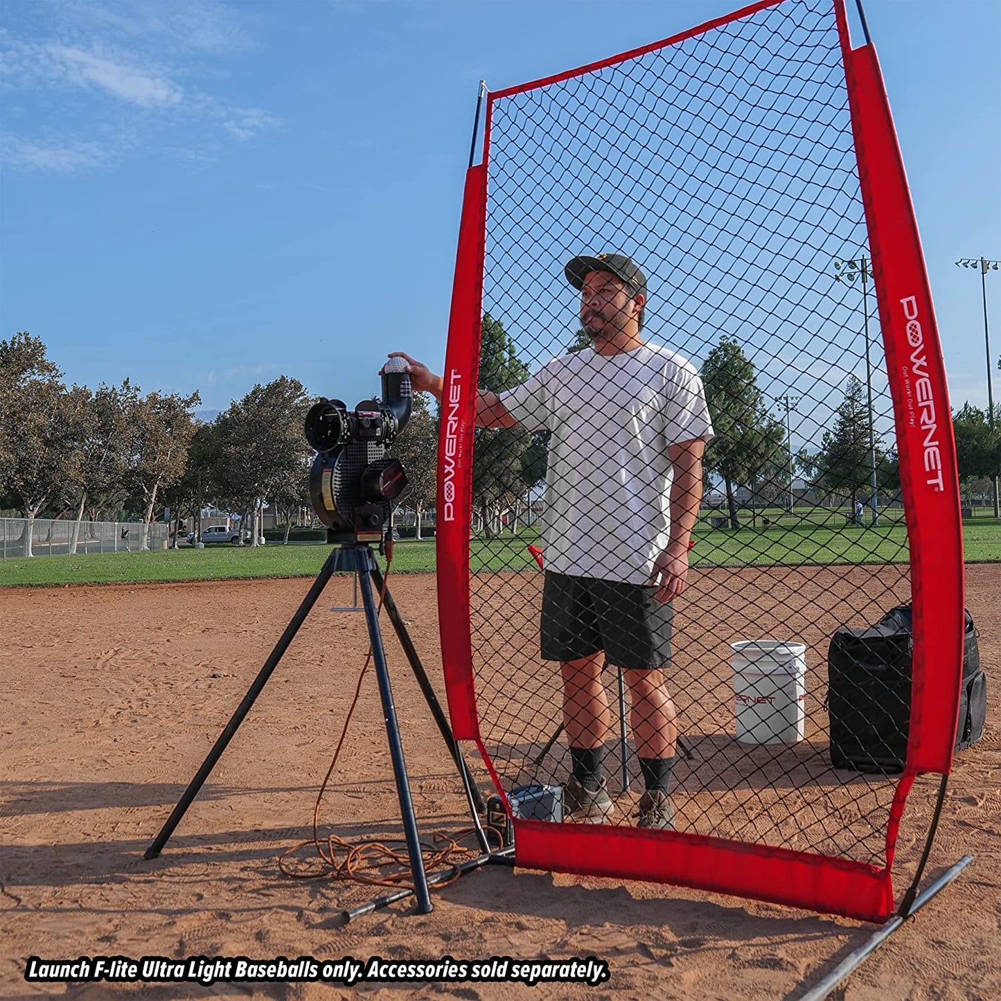 A man operates a PowerNet Launch F-Lite Pitching Machine behind a net on a baseball field, with trees and players in view. Use only PowerNet Launch F-Lite Pitching Machine Baseballs: 1194-1. Accessories sold separately.