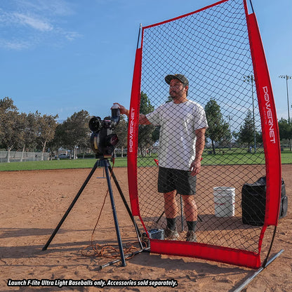 A man operates a PowerNet Launch F-Lite Pitching Machine behind a net on a baseball field, with trees and players in view. Use only PowerNet Launch F-Lite Pitching Machine Baseballs: 1194-1. Accessories sold separately.