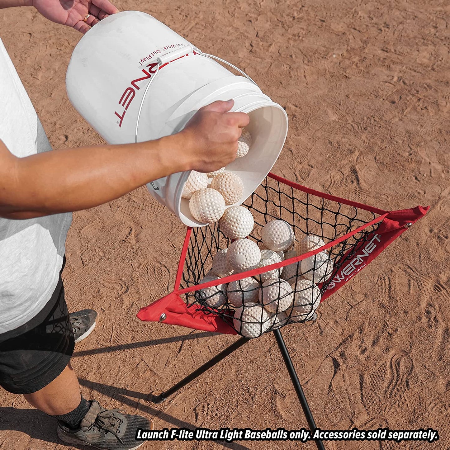 A person pours PowerNet Launch F-Lite Pitching Machine Baseballs (1194-1) into a netted basket on a tripod stand, set on a dirt field. Text reads: Use only PowerNet Launch F-Lite balls. Accessories sold separately.