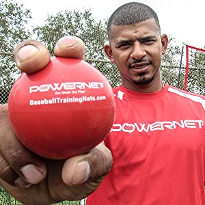 A man in a red PowerNet shirt holds a PowerNet 3.2" Weighted Hitting and Batting Training Ball (6 Pack): 1004L toward the camera, with the brand’s website on the ball, and a fence and trees visible in the background.