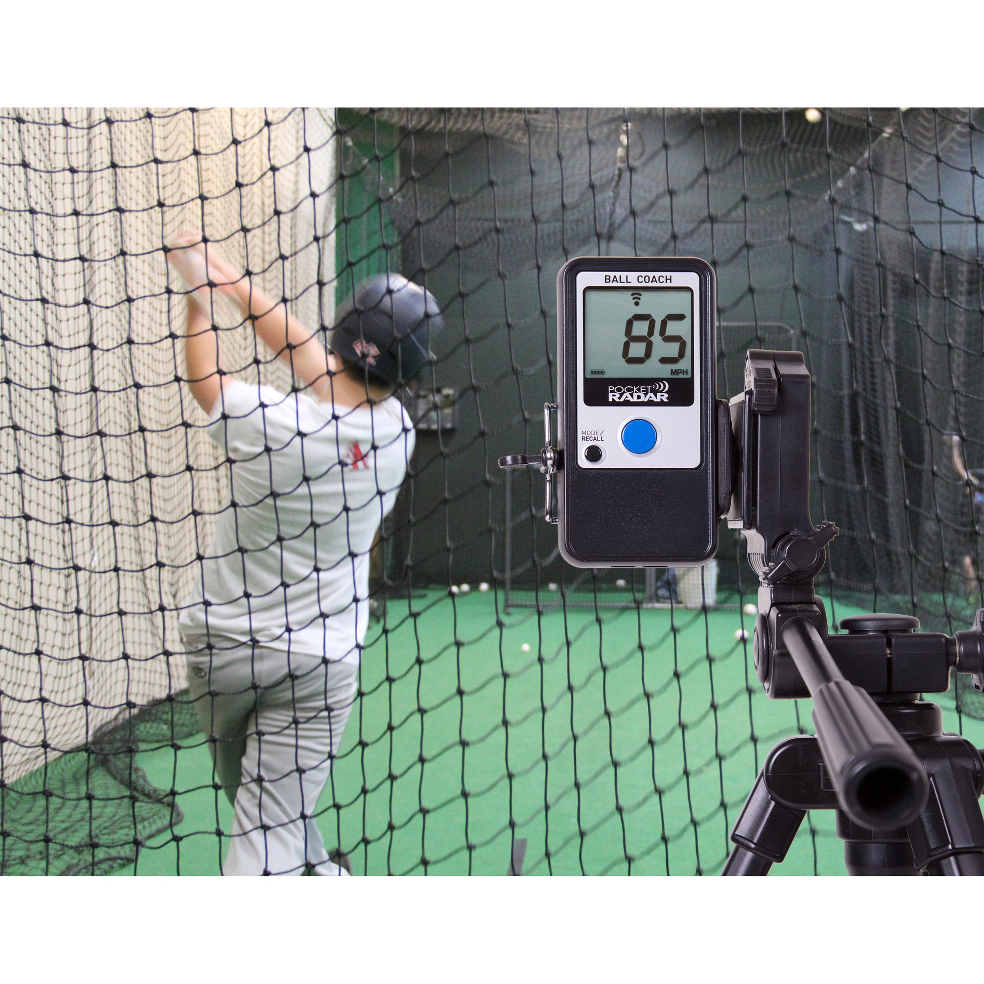 A baseball player swings in a cage as the Pocket Radar Ball Coach Radar (PR1000-BC) by Pocket Radar, set up behind a net, displays an 85 mph ball speed—providing accurate and essential speed measurements for training.