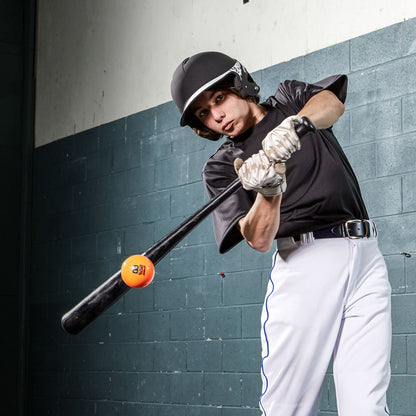 A baseball player in a black helmet and jersey and white pants swings a black bat to hit a Champro Sports 9" Control Flight Hitting Ball (CBB92) indoors against a blue and white brick wall.