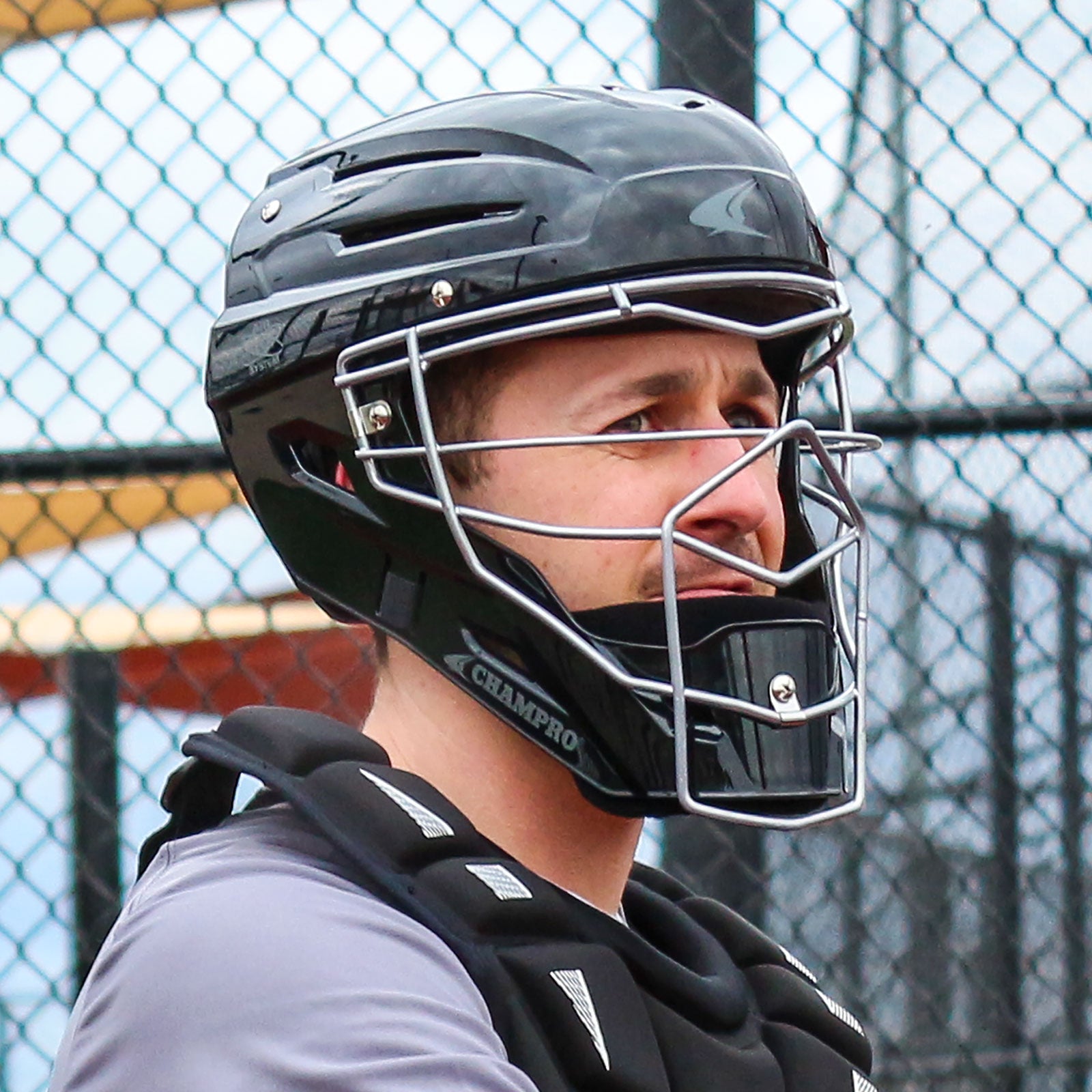 A baseball catcher in a Champro Sports Champro Optimus MVP CM75 helmet with an ABS shell and advanced ventilation system stands by a chain-link fence, looking to the side.