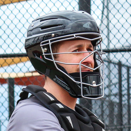 A baseball catcher in a Champro Sports Champro Optimus MVP CM75 helmet with an ABS shell and advanced ventilation system stands by a chain-link fence, looking to the side.