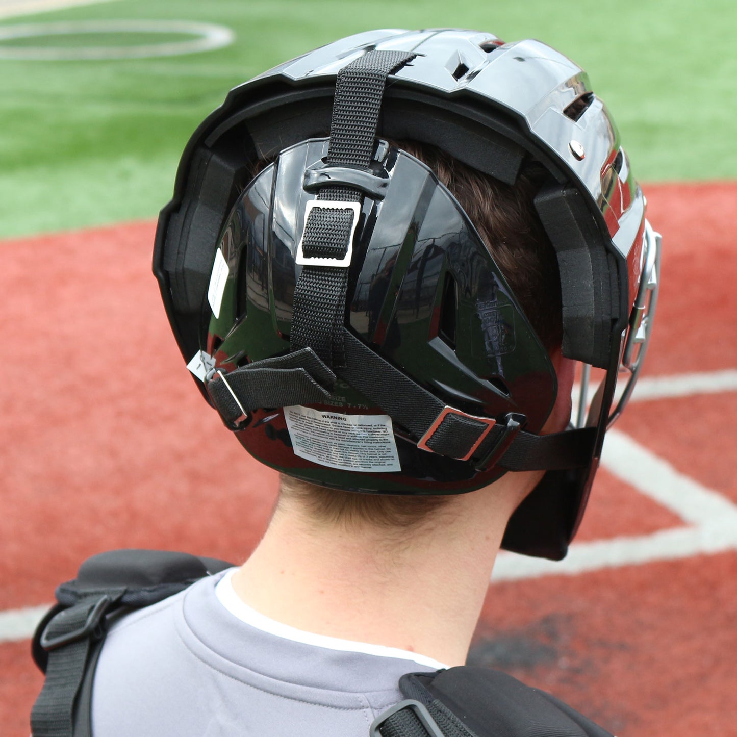 A baseball catcher wearing a Champro Sports Optimus MVP Hockey Style Catcher's Helmet (CM75) stands on red and green artificial turf, viewed from behind in shiny black gear with ventilation system and chest protection.
