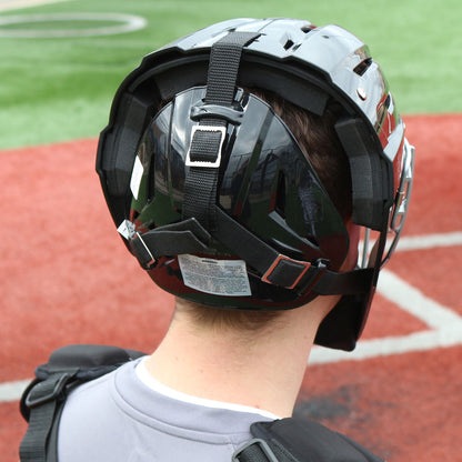 A baseball catcher wearing a Champro Sports Optimus MVP Hockey Style Catcher's Helmet (CM75) stands on red and green artificial turf, viewed from behind in shiny black gear with ventilation system and chest protection.