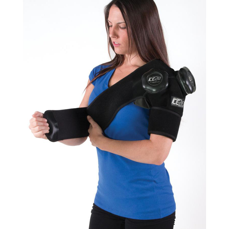 A woman in a blue shirt wears a Bownet ICE20 Double Shoulder Ice Compression Wrap (ICE-Dbl-Shoulder), adjusting the arm strap with her left hand to secure two ice packs for targeted therapy.