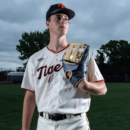 A young baseball player in a white Tigers uniform stands on the field, confidently holding a Rawlings REV1X 12.75" Baseball Glove (REV3039-6) as trees and a cloudy sky form the backdrop.