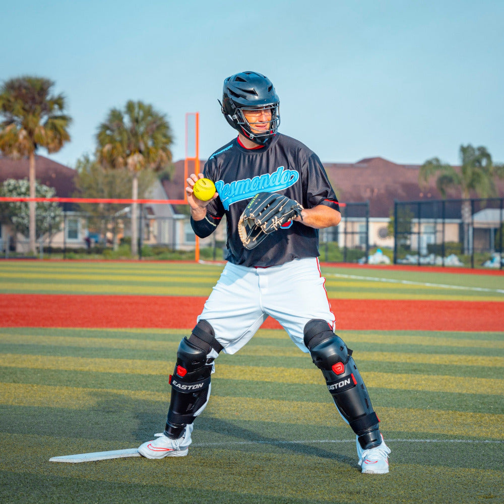 A baseball catcher wearing an Easton Hellcat Slowpitch Fielding Helmet (EHCATH) by Easton stands on the field, holding a yellow ball and glove, ready to throw. Trees and buildings are visible in the background.