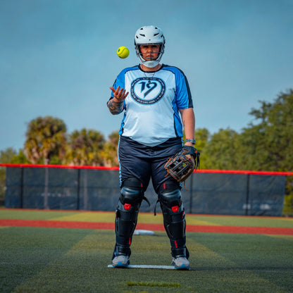 A softball player wearing an Easton Hellcat Slowpitch Fielding Helmet (EHCATH) by Easton stands on the field with catcher’s gear, tossing a softball. Trees and a red fence appear in the background under a clear sky.