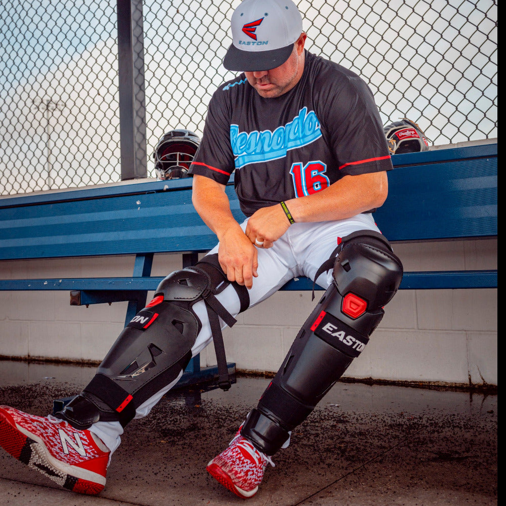 A baseball player in uniform sits on a blue bench by a chain-link fence, lacing up red sneakers and putting on Easton Hellcat Slowpitch Fielding Leg Guards (EHCATL) by Easton. A helmet and gloves lie beside him.